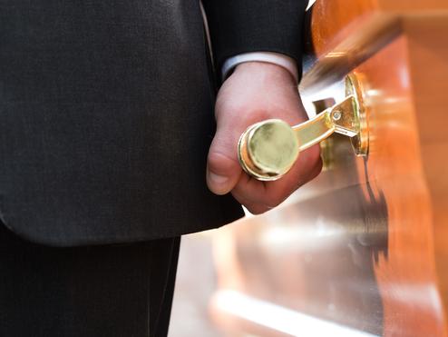 Mann in Anzug hält eine goldene Griffschleife an einem Holzsarg.